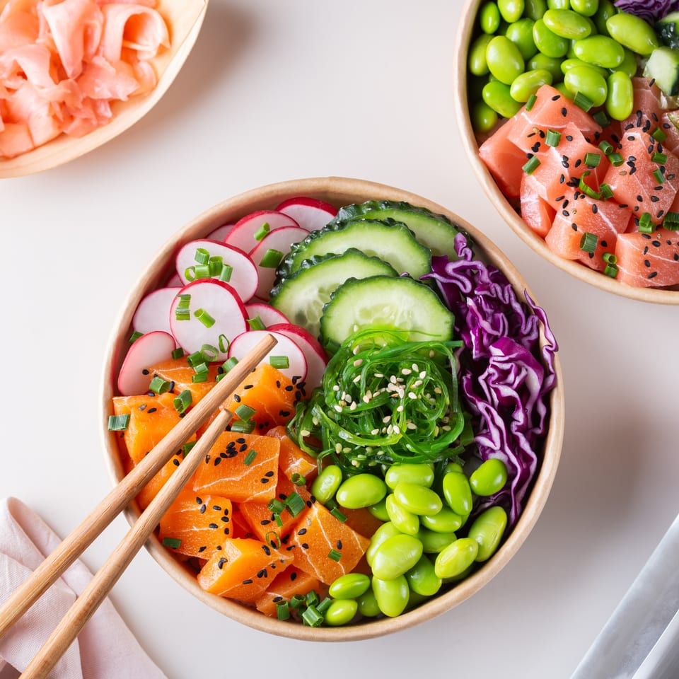 Two colorful poke bowls with salmon, edamame, seaweed, and vegetables, with chopsticks on a white table.