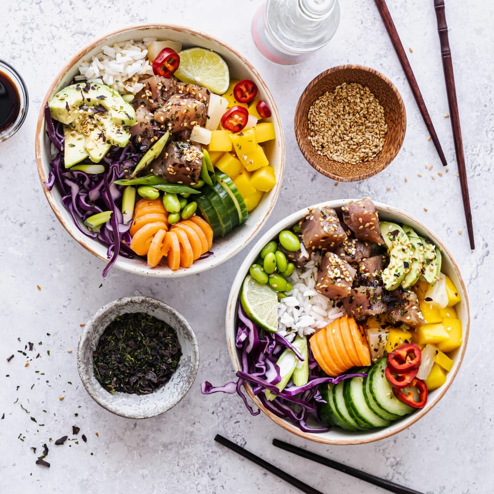 Two colorful poke bowls with tuna, rice, avocado, mango, and vegetables, with chopsticks and seasonings.
